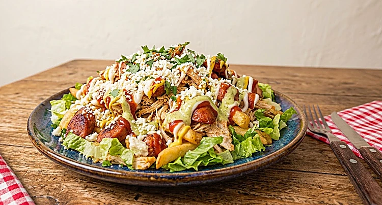 Salchipapa servida en plato azul con salchicha, papas, lechuga, queso y salsas, sobre mesa de madera